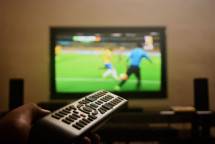 Close-up of a hand holding a remote control in front of a TV showing a football match. (Foto: Soumith Soman / Pexels)